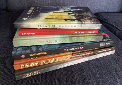 A staggered pile of hardback books, showing spines and part of the front covers. From bottom to top: 'Conan the Barbarian' for Modiphius Conan; 'Shadows Over Stillwater' for Call of Cthulhu; 'The Horned Rat' for Warhammer Fantasy; 'Out of the Ashes' corebook; 'Sanction' corebook; and 'Liminal Casebook'.